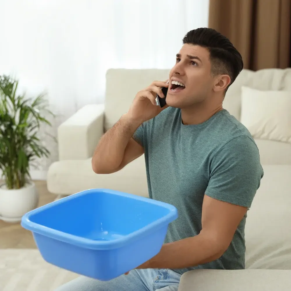 A homeowner holds a blue plastic bucket to catch dripping water whilst on the phone arranging Emergency Leak Repairs to his home